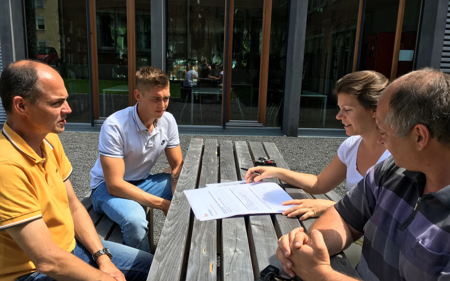 Vier Personen, zwei Männer, eine Frau und ein junger Mann, sitzen an einem Holztisch im Freien und besprechen Dokumente (Workshop).