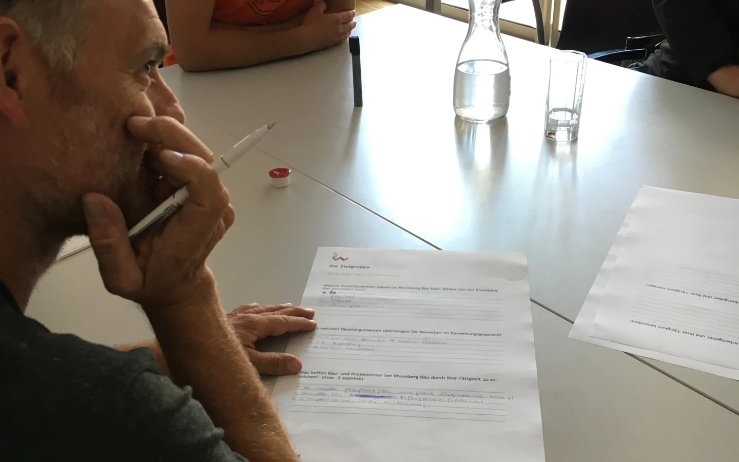 Zwei Männer sitzen an einem Tisch, auf dem Papiere, Stifte und eine Kaffeetasse liegen. Im Hintergrund ist ein Architekturmodell sichtbar (Workshop).