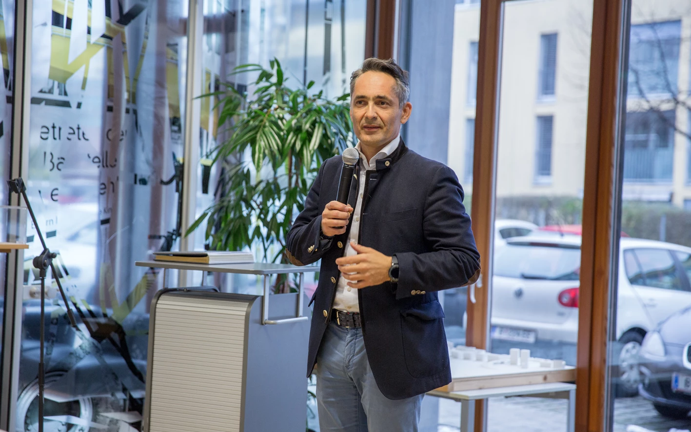 Ein Mann im Sakko spricht vor einem Mikrofon in einem modernen Raum mit Glasfenstern und Ausblick ins Freie (Hubert, Geburtstag).