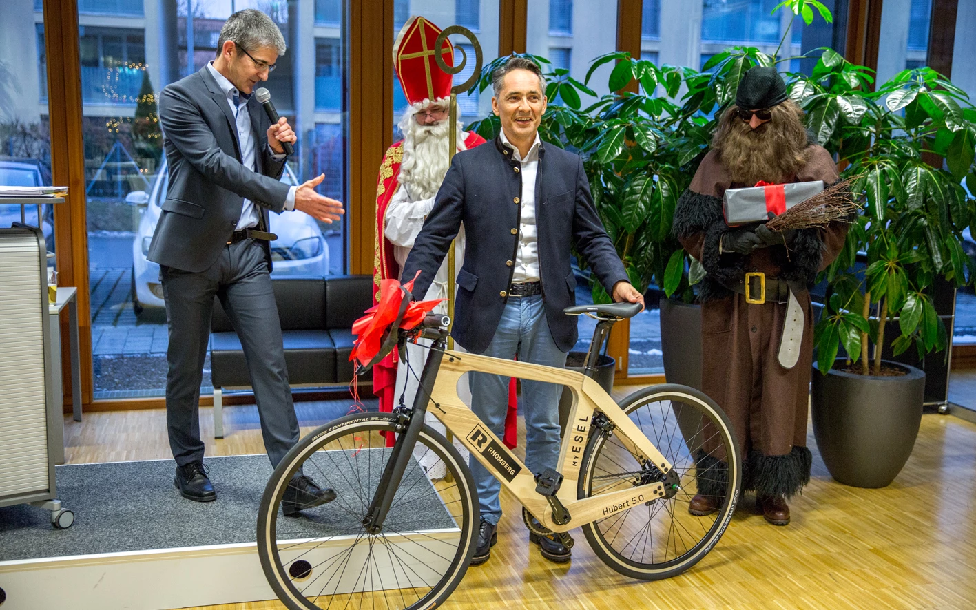 Geschäftsleute und verkleidete Personen präsentieren ein Holzfahrrad Hubert 5.0 mit dem Rhomberg Logo auf einer Bühne (Geburtstag).