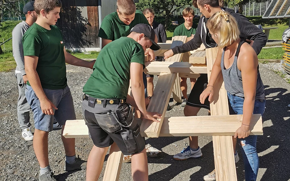 Mehrere Lehrlinge von Rhomberg Bau montieren gemeinsam an einem sonnigen Tag Holzteile im Freien (Zusammenbauen).