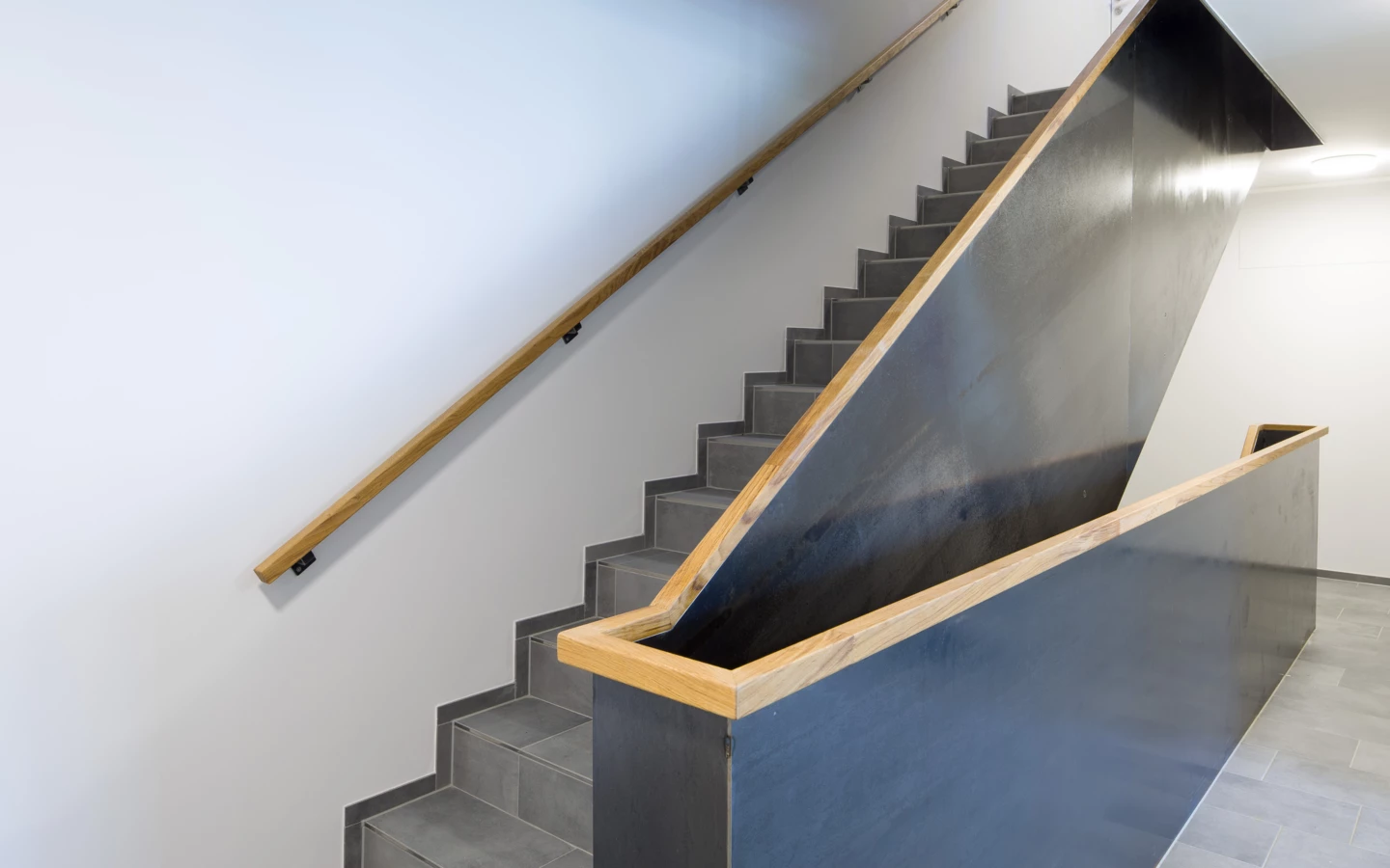 Eine moderne Treppe mit Holzgeländern und dunkelgrauen Metallbrüstungen in einem Gebäude der Unterhochstegstrasse
