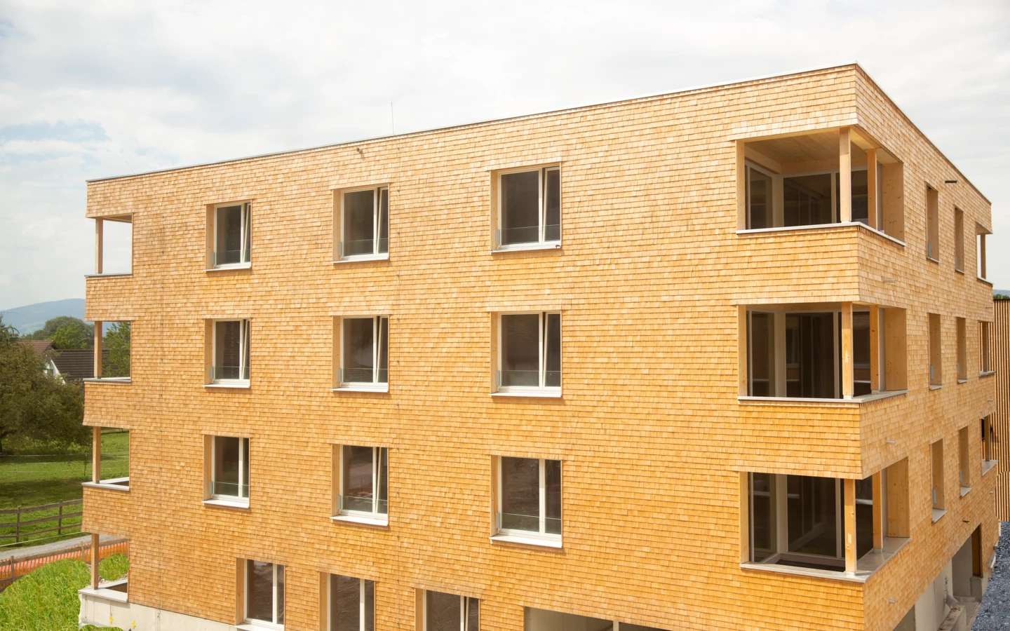 Modernes Mehrfamilienhaus mit Holzfassade und vielen Fenstern, umgeben von grüner Landschaft und Bäumen unter bewölktem Himmel (Hellbrunnenstraße).