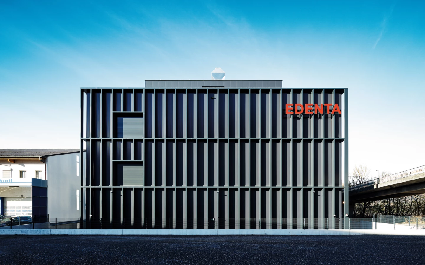Modernes, quaderförmiges Geschäftsgebäude mit vertikalen Lamellen und Leuchtschrift „EDENTA“ an der Fassade unter blauem Himmel.