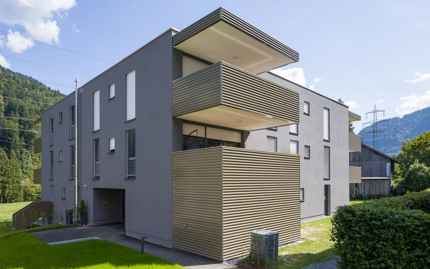 Moderne Wohnanlage in Vandans mit grauer Fassade, Balkonen mit Holzlamellen und grünem Rasen im Vordergrund unter blauem Himmel (Bahnhofstraße, Bahnhofsstrasse)
