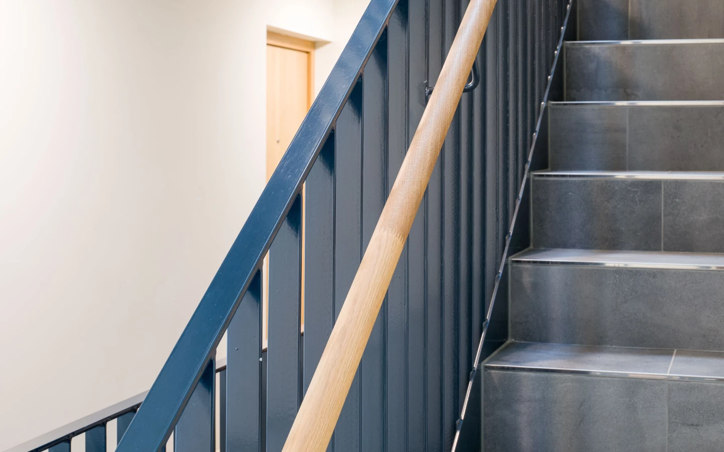 Eine dunkelblaue Metalltreppe mit einem Holzhandlauf und grau gefliesten Stufen in der Wohnanlage Bahnhofsstrasse Vandans.