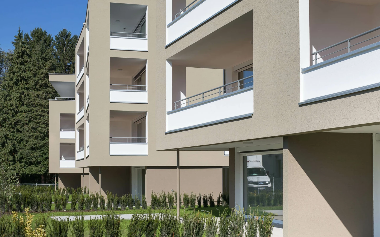 Moderne Wohnanlage Widum Lustenau mit hellgrauer Fassade, weissen Balkonbrüstungen und gepflegter Grünfläche mit Büschen