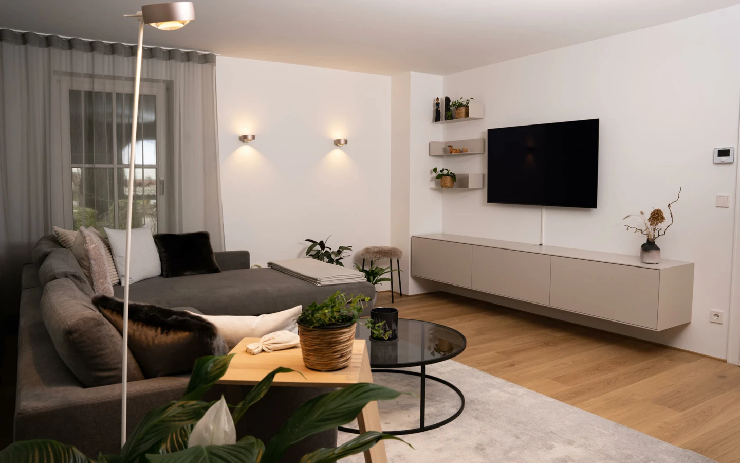 Modernes Wohnzimmer mit grauem Ecksofa, hellem Holzboden, TV, Wandleuchten und Zimmerpflanzen (Generalsanierung, Bohle)