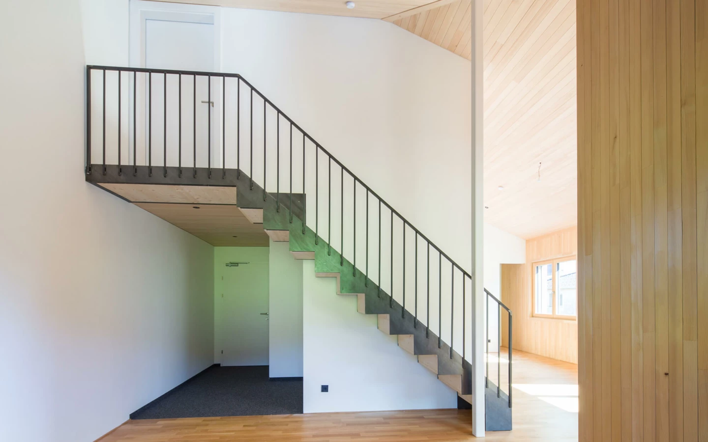 Eine moderne Holztreppe mit schwarzem Metallgeländer in einem hellen Eingangsbereich mit Holzboden und weißen Wänden (Wohnanlage, Alberschwende).