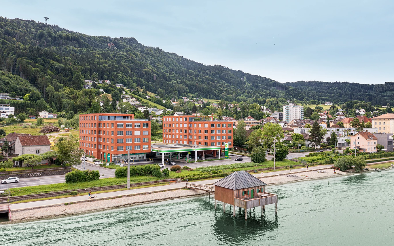 Zwei moderne rote Backsteingebäude neben einer BP-Tankstelle an einer Straße am Bodensee. Ein Holzhaus auf Stelzen ragt ins Wasser, dahinter ein bewaldeter Berg (Seeblick, Lochau).