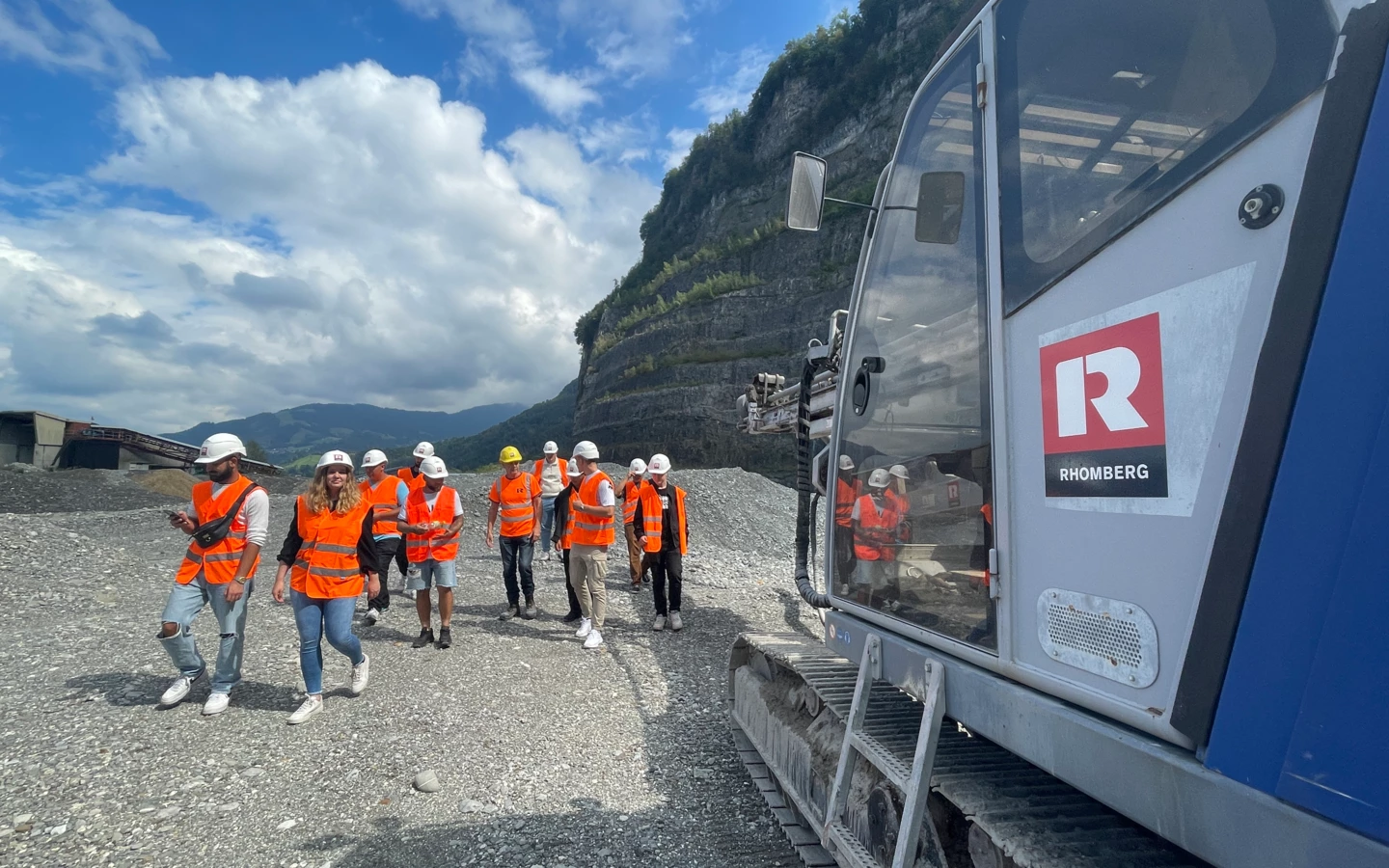 Eine Gruppe junger Menschen in orangefarbenen Westen und Schutzhelmen geht an einem Bagger mit Rhomberg-Logo vorbei auf einem Kiesweg, mit Bergen im Hintergrund (Lehre).