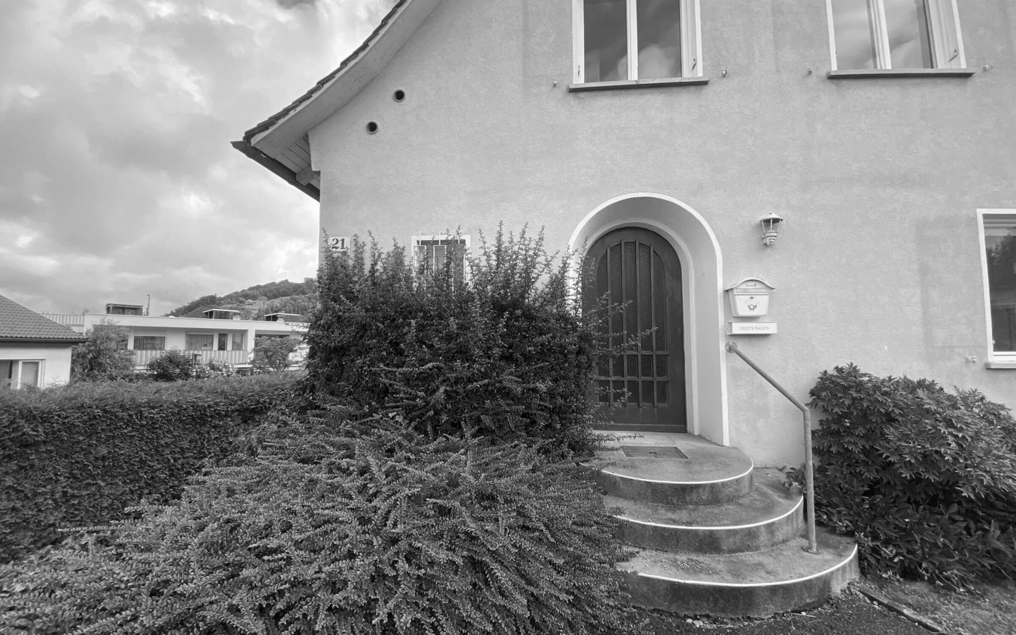 Schwarz-Weiß-Aufnahme des Eingangs eines Einfamilienhauses in Dornbirn mit Büschen und runden Treppen vor der Sanierung (Generalsanierung).