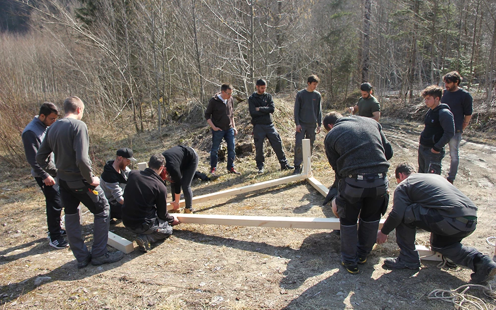 Eine Gruppe junger Männer arbeitet im Freien an einer Holzkonstruktion, umgeben von Bäumen und Sträuchern (Lehrlingstag).