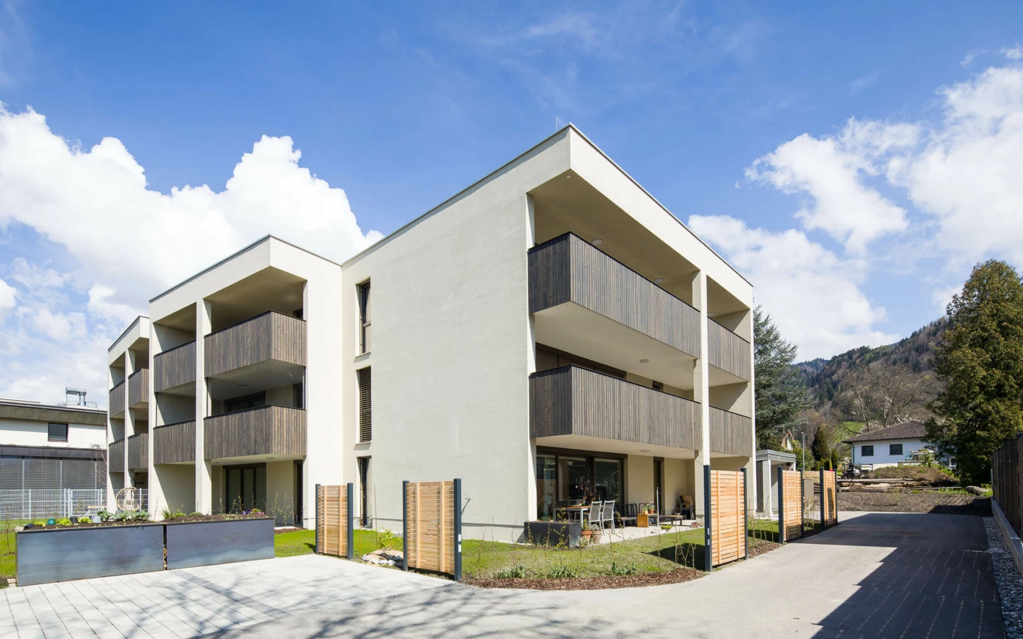 Modernes Mehrfamilienhaus mit hellbeiger Fassade und Holzbalkonen in Dornbirn, unter blauem Himmel mit weißen Wolken (Feldgasse, Wohnanlage).