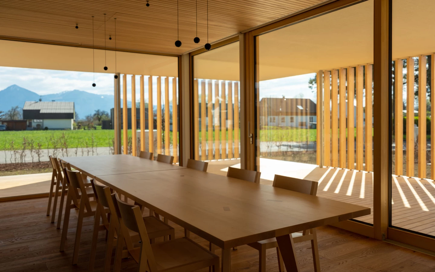 Ein großer Holztisch mit Holzstühlen in einem modernen Raum mit Holzvertäfelung und großen Fenstern, die einen Blick auf Berge und die grüne Landschaft bieten (Wolfurt, Dieter).