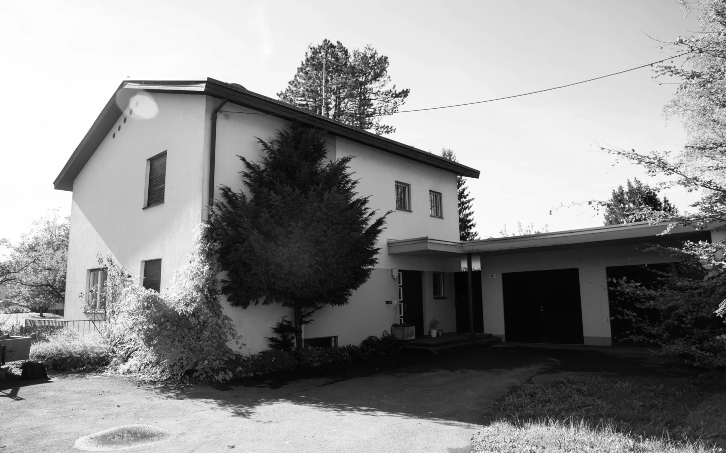 Schwarz-Weiß-Aufnahme eines alten Einfamilienhauses in Dornbirn mit Garagen und Carport, umgeben von Bäumen und Sträuchern (Generalsanierung, Holzoptik).