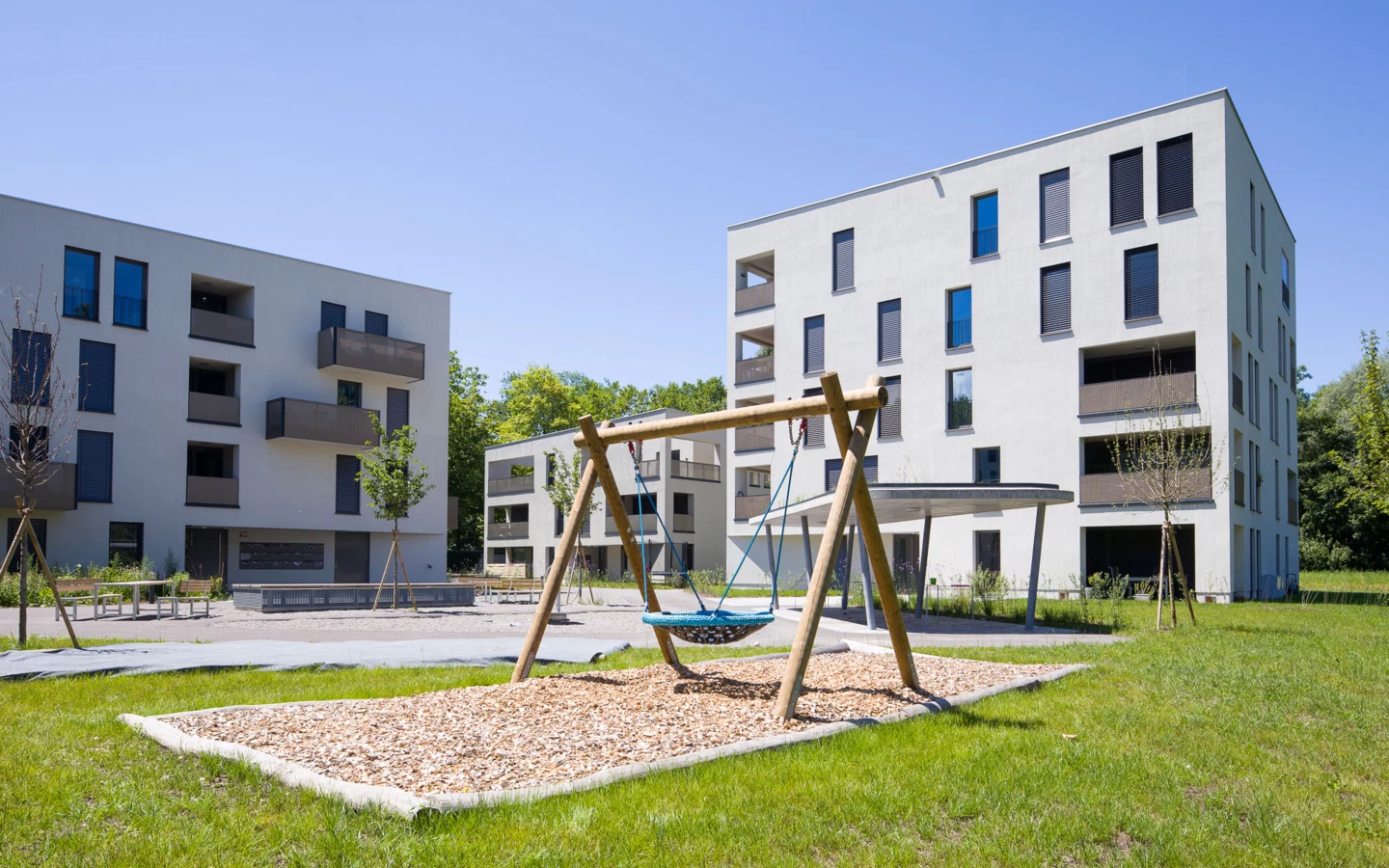 Moderne Wohnanlage 'Wohnen an der Furt' mit weiß gestrichenen Fassaden, dunklen Balkonen und großen Fenstern, im Vordergrund ein Kinderspielplatz mit Schaukel (Eigentumswohnung).