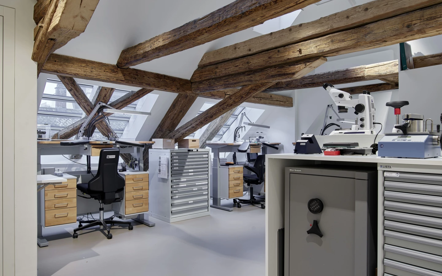 Uhrmacherwerkstatt unter dem Dachgiebel mit Holzbalken. Mehrere Arbeitsplätze mit Mikroskopen, Werkzeugen und Lagerregalen (Huber, Rathausstr).