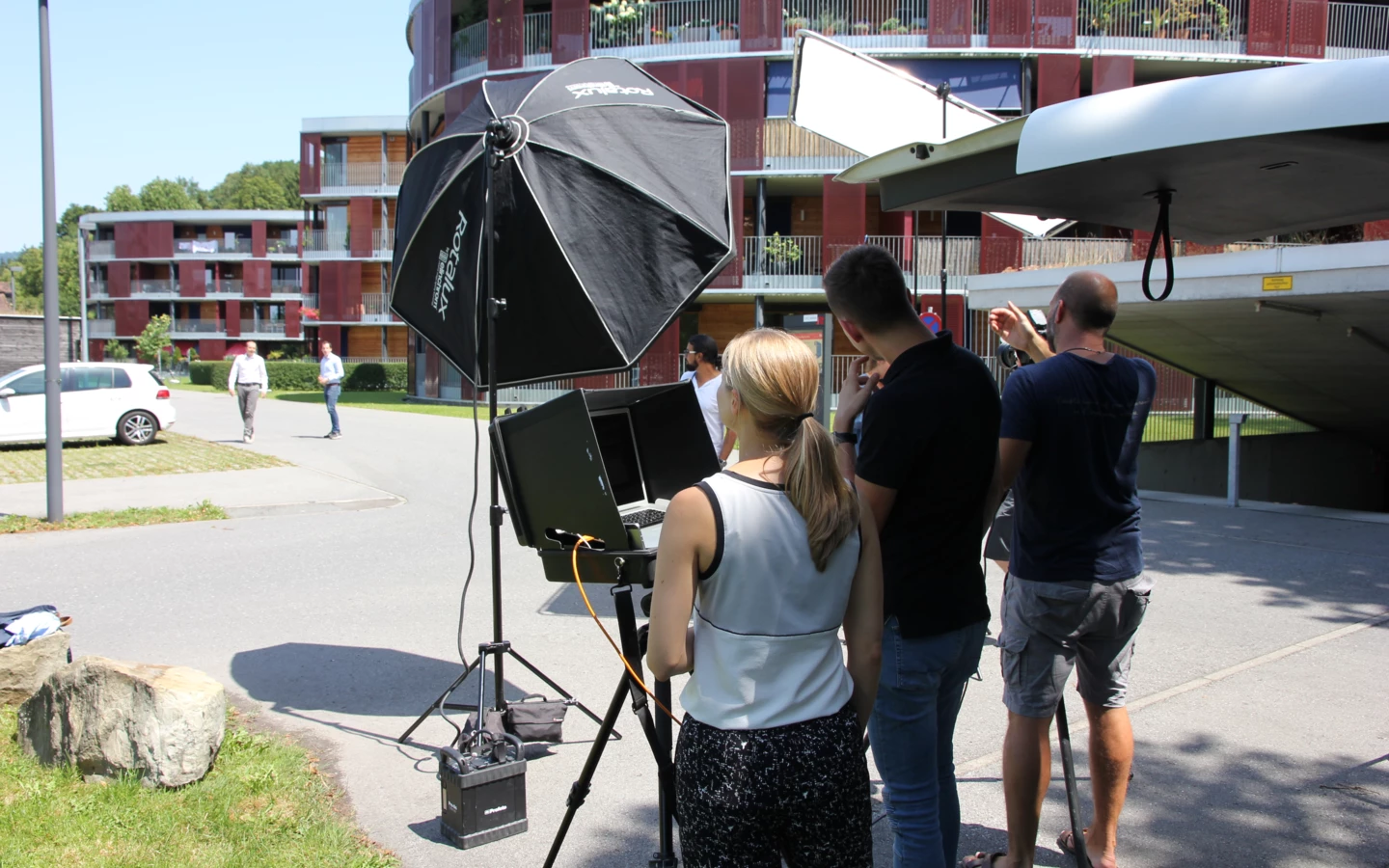 Team bei einem Fotoshooting im Freien, mit Kameraausrüstung, Stativen und Softboxen vor einem modernen Gebäude.