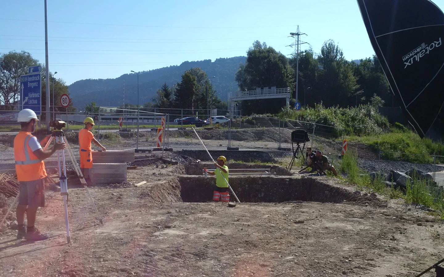 Rhomberg Bau Mitarbeiter auf einer Baustelle mit Vermessungsgeräten und Kamera-Equipment unter blauem Himmel.