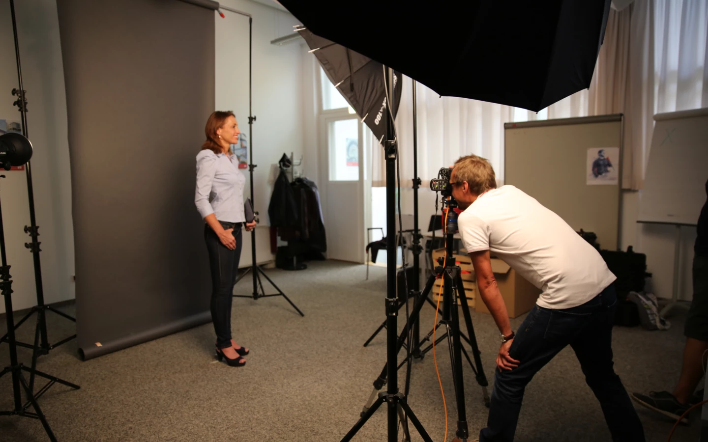 Ein Fotograf macht ein Porträtfoto einer Frau in einem Fotoshooting-Studio mit grauer Kulisse und Beleuchtungsausrüstung.