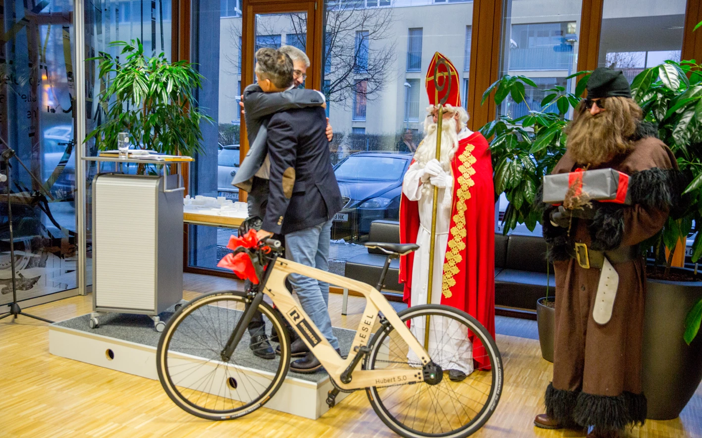 Zwei Männer umarmen sich neben einem hölzernen Rhomberg Fahrrad, während Nikolaus und Krampus bereitstehen (Hubert, Geburtstag).