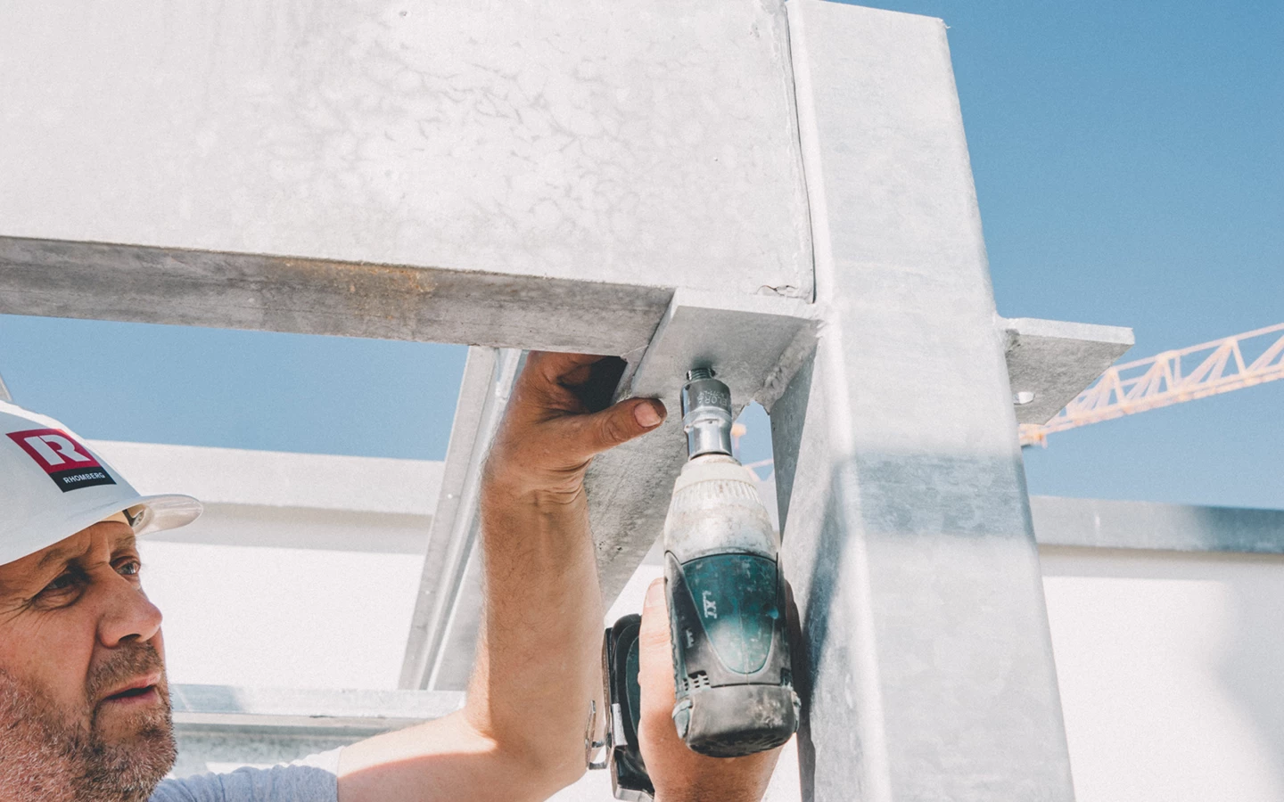 Ein Bauarbeiter in einem weißen Helm mit rotem Rhomberg-Logo schraubt ein Metallteil an einer Baustelle fest, während die Sonne scheint (Hochbau).