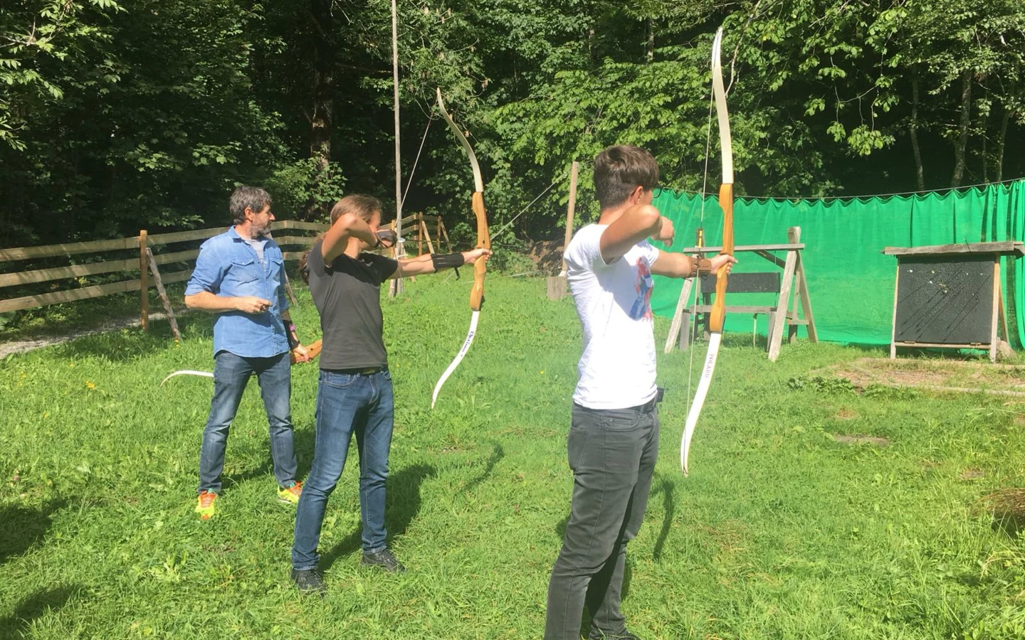 Drei Personen, vermutlich Lehrlinge, beim Bogenschiessen im Freien, konzentriert auf das Ziel.