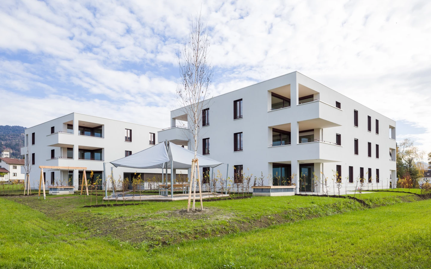 Zwei moderne weiße Wohngebäude mit Balkonen, umgeben von grünen Rasenflächen, mit einem überdachten Sitzbereich und jungen Bäumen (Unterhochstegstrasse).