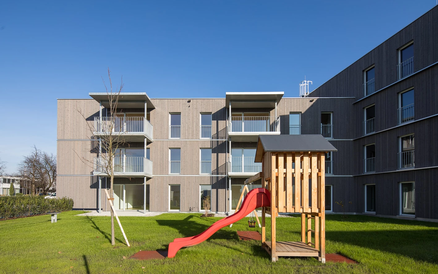 Modernes Mehrfamilienhaus mit Holzfassade, Balkonen und einem Spielplatz mit roter Rutsche bei sonnigem Wetter (Froschweg).