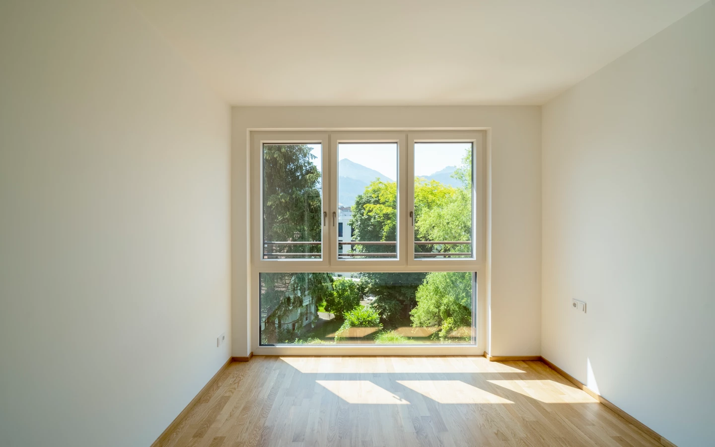 Leeres Zimmer mit Holzboden und einem großen Fenster, das auf grüne Bäume und Berge in Dornbirn blickt