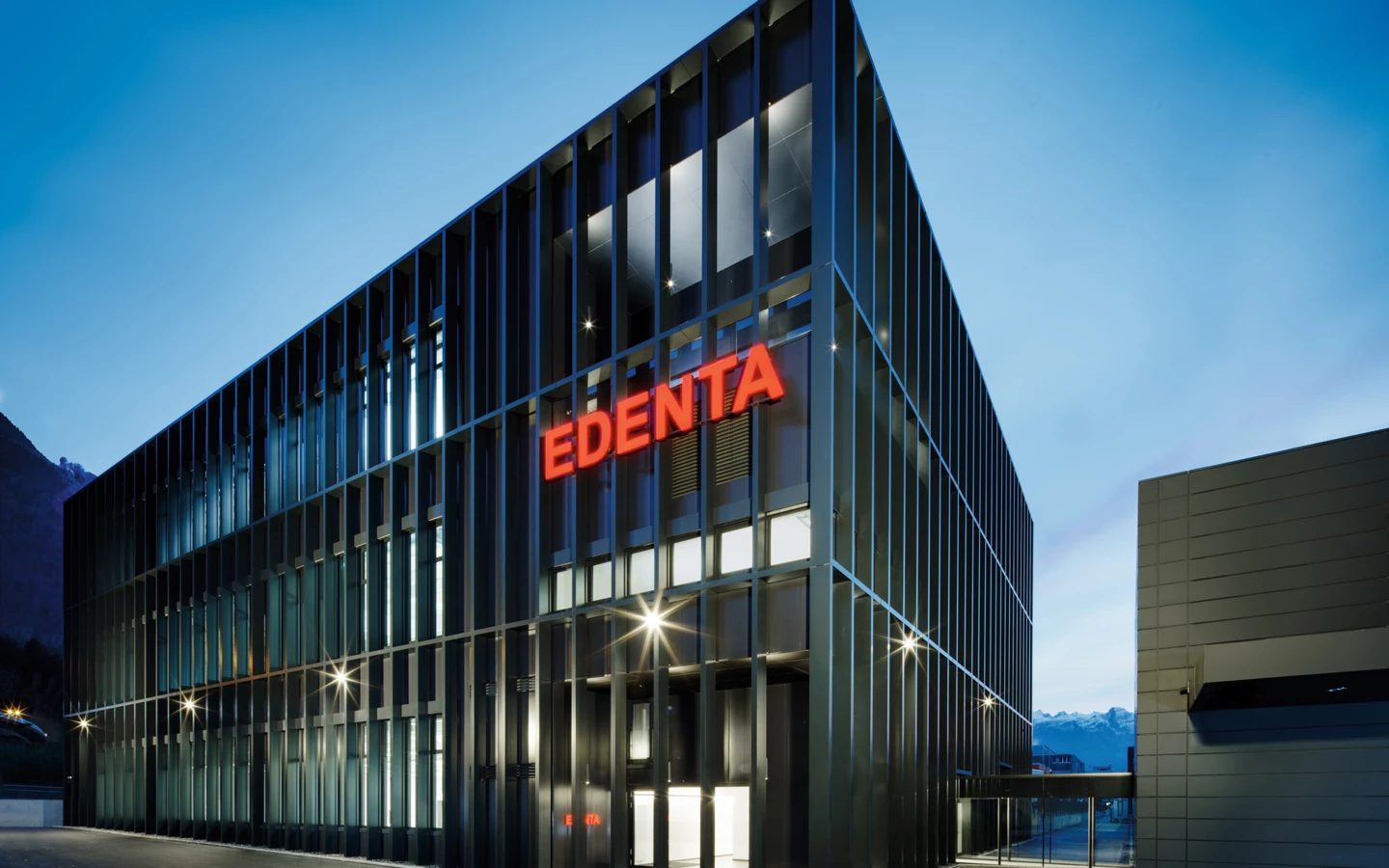 Modernes Geschäftsgebäude von Edenta mit horizontalen und vertikalen Lamellen, beleuchtet in der Dämmerung unter blauem Himmel.