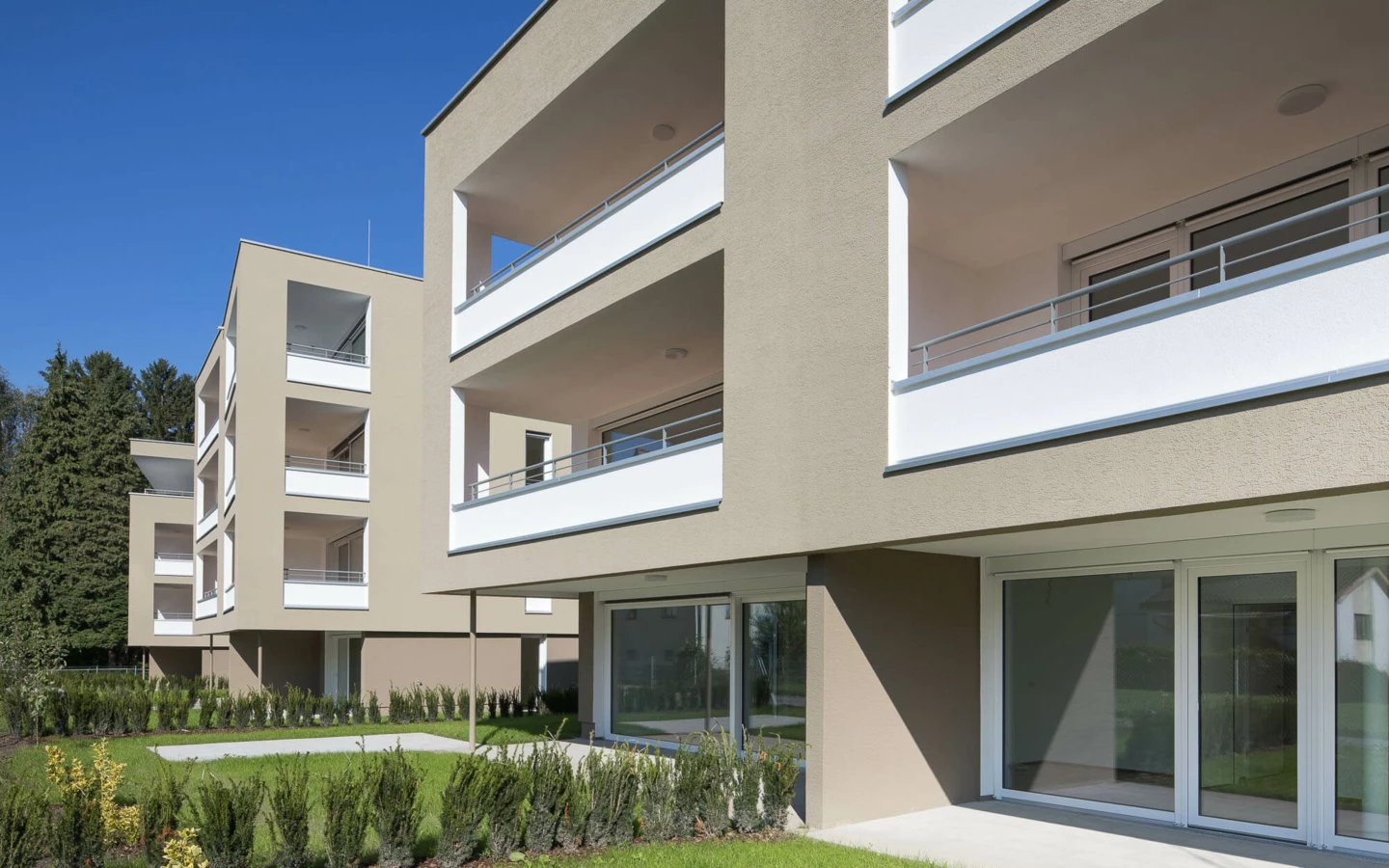 Moderne, hellbraune Wohngebäude mit weißen Balkonen und großen Fenstern, umgeben von grünen Rasenflächen und Hecken unter blauem Himmel in der Wohnanlage Widum Lustenau.