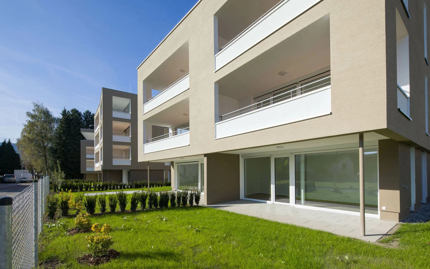 Moderne Wohnanlage Widum in Lustenau mit mehreren Gebäuden, gepflegten Rasenflächen und Hecken unter blauem Himmel.