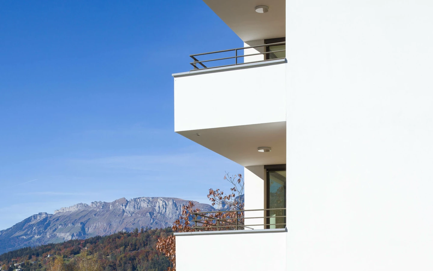 Moderne weiße Balkone eines Mehrfamilienhauses mit Bergblick an der Wolf-Huber-Straße in Feldkirch unter blauem Himmel (Wohnanlage).