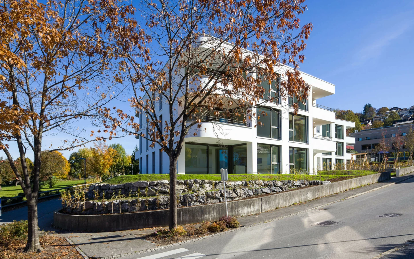 Moderne weiße Wohnanlage an der Wolf-Huber-Straße in Feldkirch unter blauem Himmel, mit Herbstbäumen und Rasenfläche.