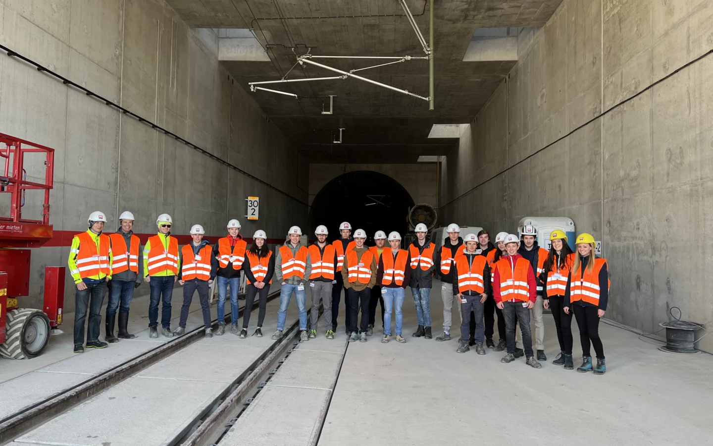Eine Gruppe von 18 Personen in Sicherheitskleidung mit Helmen und Warnwesten steht vor einem Tunnelportal mit Gleisen (Rankweil, Deutschland, Gruppenfoto).