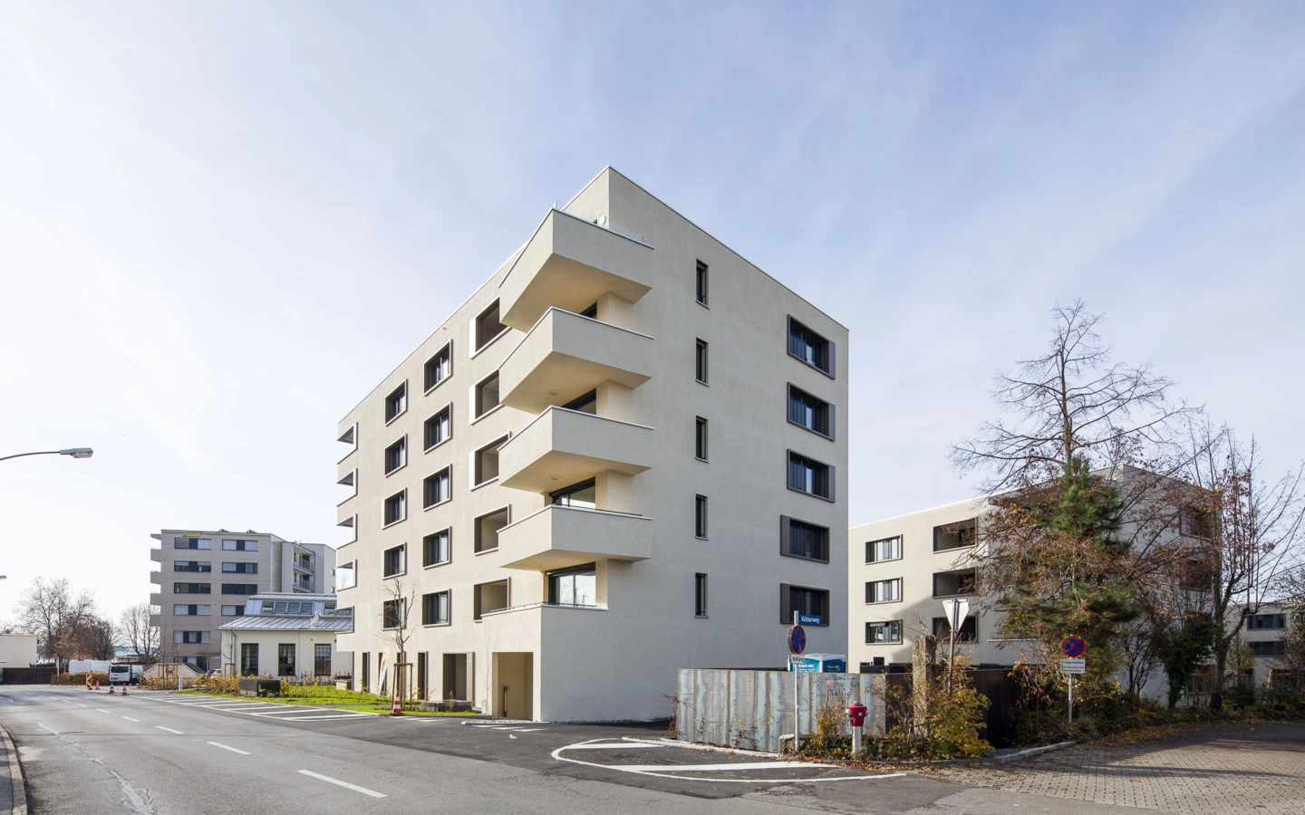 Modernes, mehrstöckiges Wohngebäude mit Balkonen an einer Straße, flankiert von Bäumen und weiteren Gebäuden unter blauem Himmel (Bäumleareal).