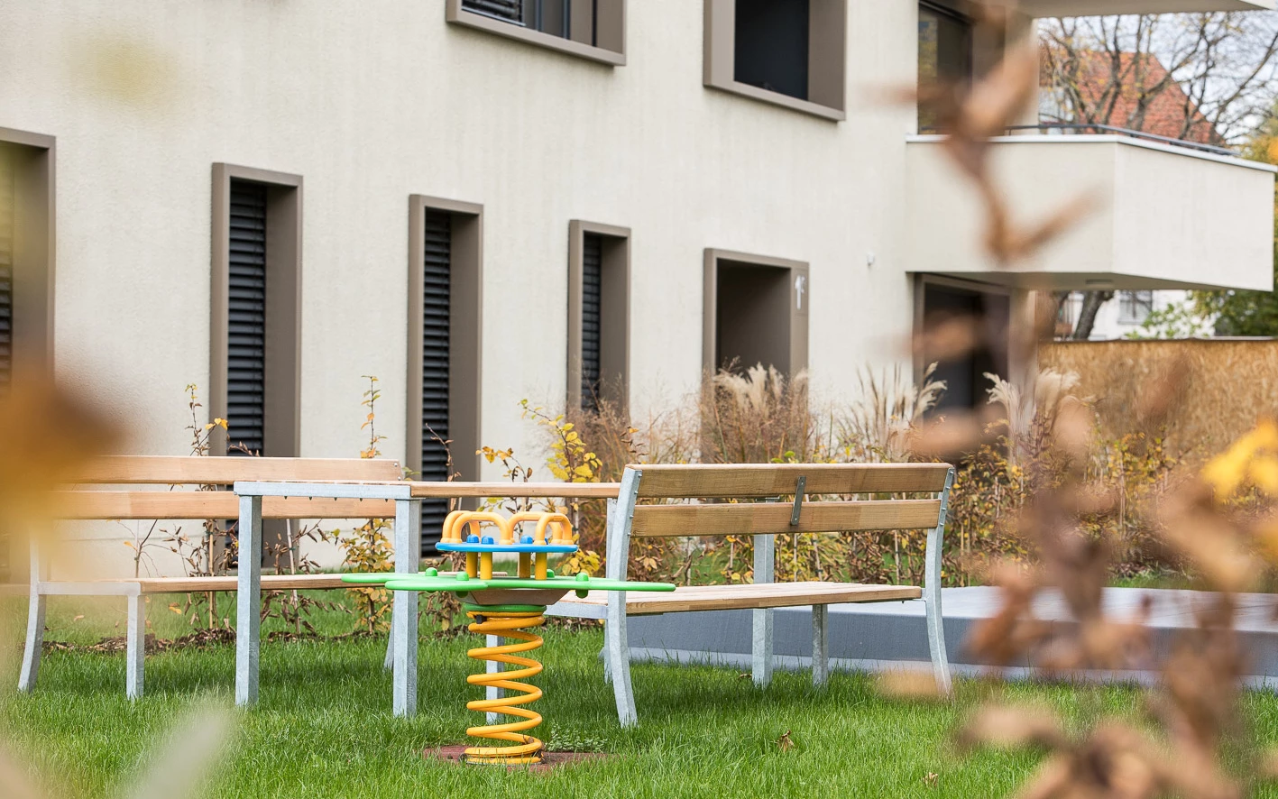 Spielplatz mit gelbem Wipptier und Holzbänken vor einem modernen Wohngebäude mit Schiebefenstern (Baeumlequartier)