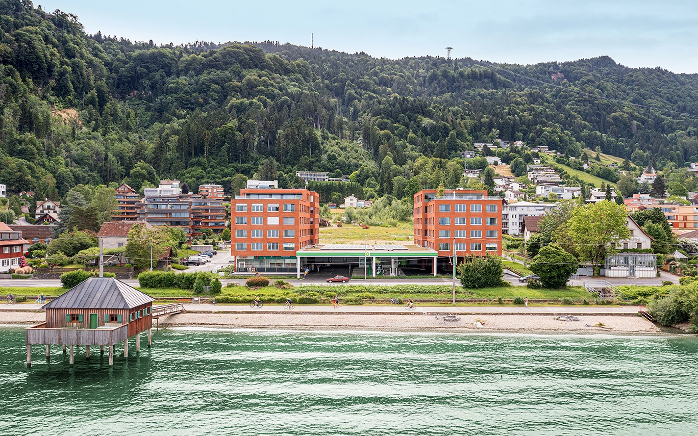 Zwei moderne rote Backsteingebäude mit einer Tankstelle dazwischen, am Ufer eines Sees mit Bergen und Bäumen im Hintergrund (Seeblick, Lochau).