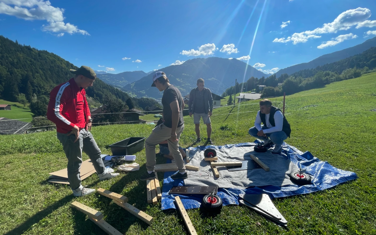 Vier junge Männer bauen im Freien auf einer blauen Plane ein Objekt zusammen, umgeben von grünen Wiesen und Bergen unter blauem Himmel mit Sonnenschein (Lehre).