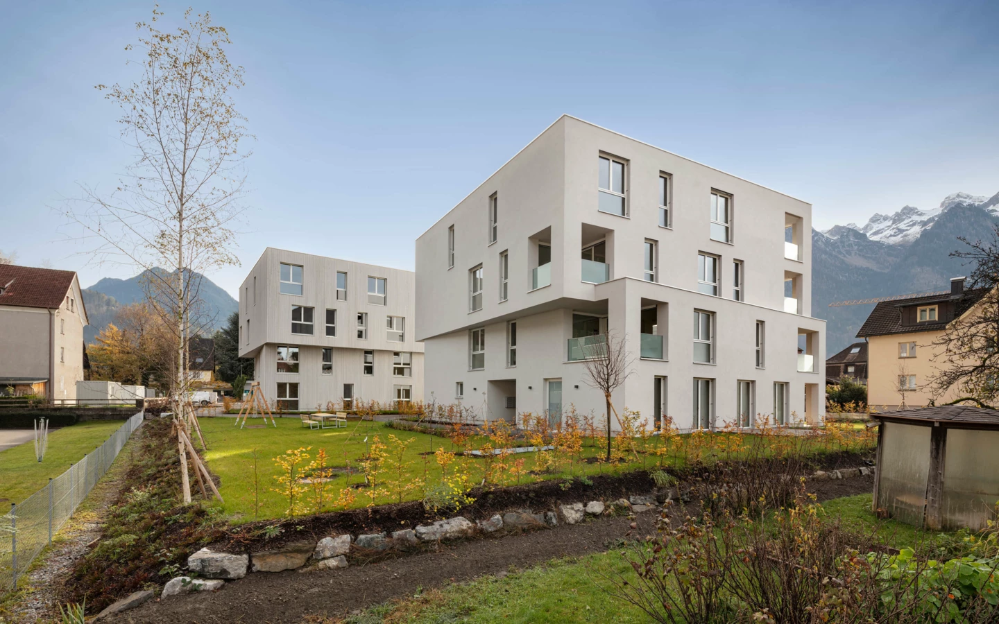 Zwei moderne, helle Mehrfamilienhäuser mit Balkonen und großem Garten auf der Wohnanlage Zürcherstrasse Bludenz vor Bergkulisse.