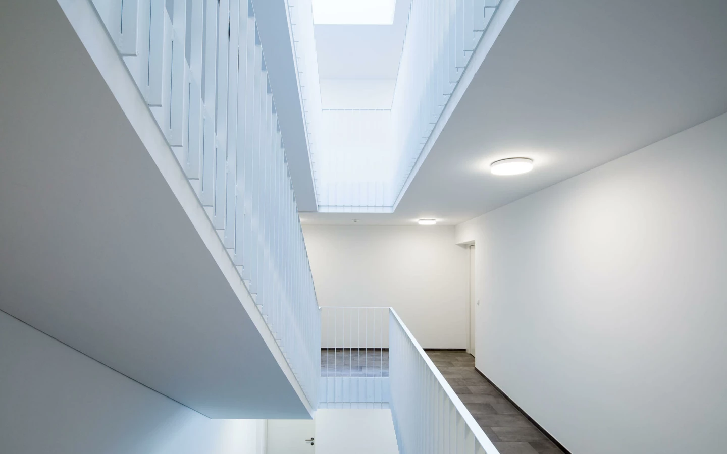 Blick von unten nach oben in ein modernes, helles Treppenhaus mit weißen Geländern, Wänden und Decken, beleuchtet durch Oberlichter sowie runde Deckenlampen (Wuhrbaumweg, Wohnanlage).