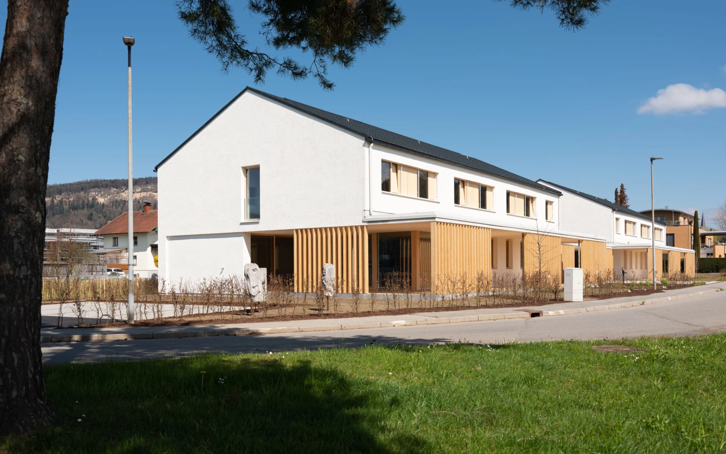 Eine Reihe moderner weißer Gebäude mit Holzfassadenelementen unter blauem Himmel, entlang einer Straße mit Grünstreifen (Wolfurt, Dieter).