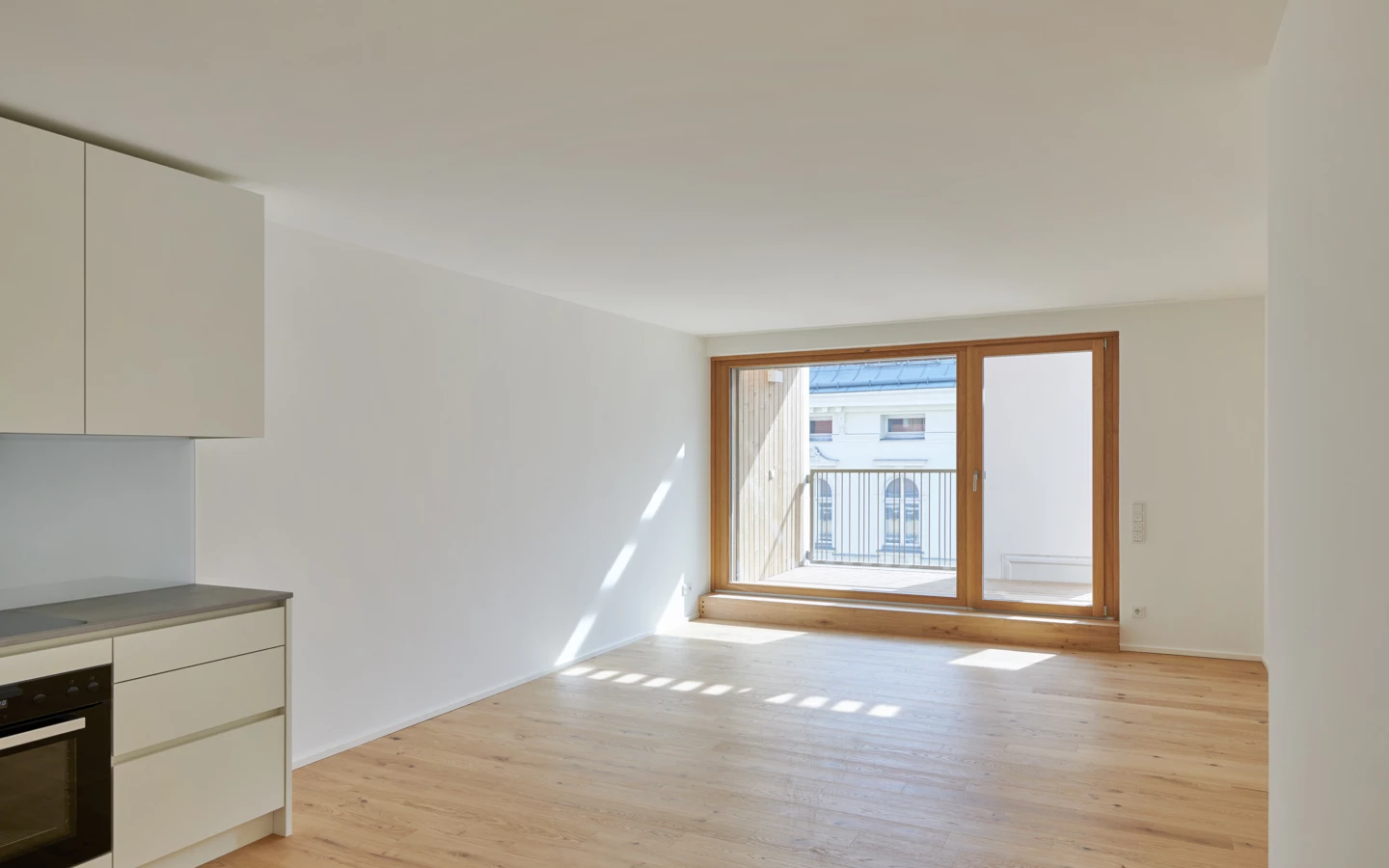 Heller Wohnraum mit Holzboden und einer modernen Küchenzeile mit Ofen und Dunstabzugshaube, Tageslicht fällt durch die Balkontür (Apollogasse).