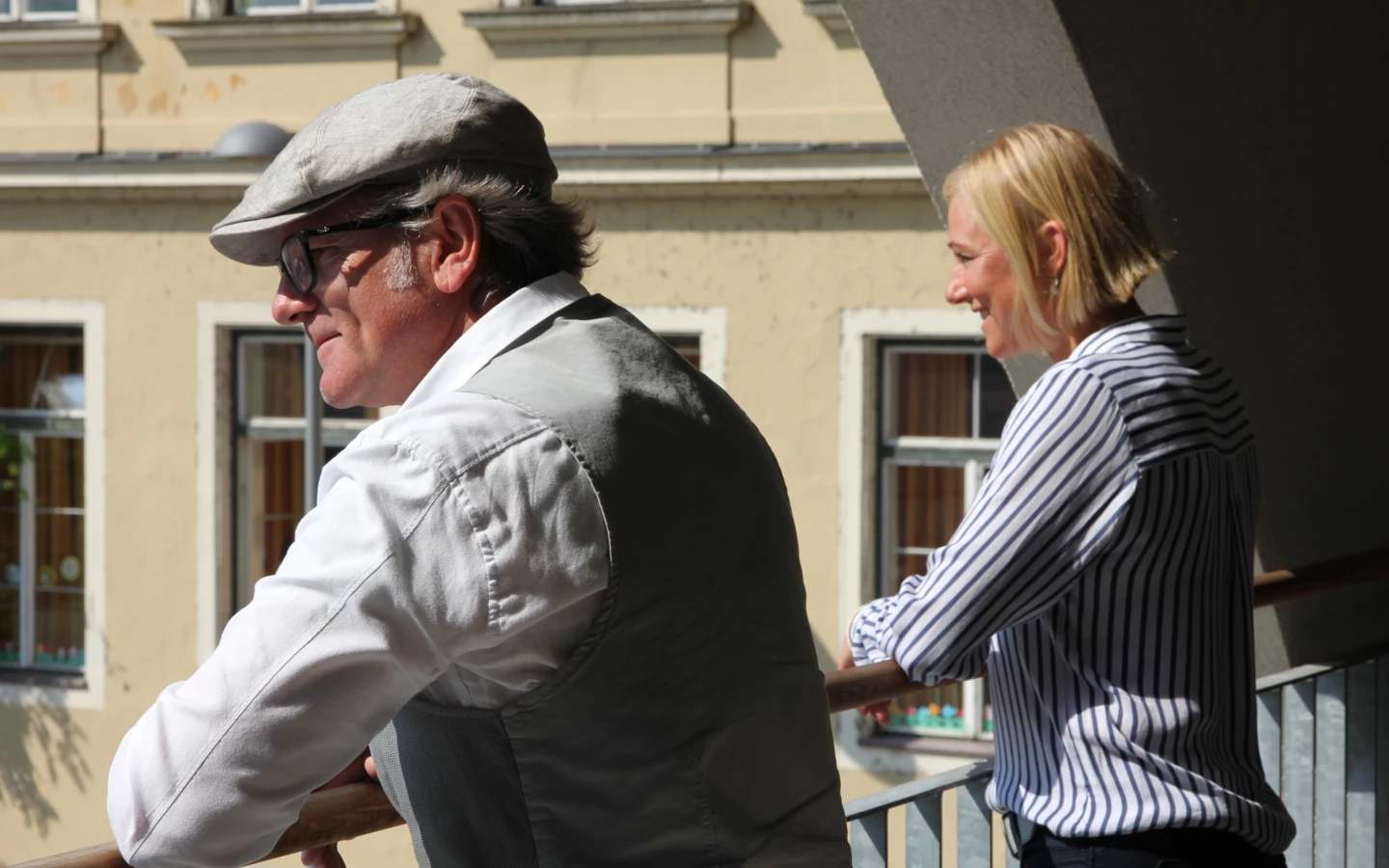 Ein Mann mit Schirmmütze und Weste und eine lachende Frau mit gestreifter Bluse stehen auf einem Balkon.