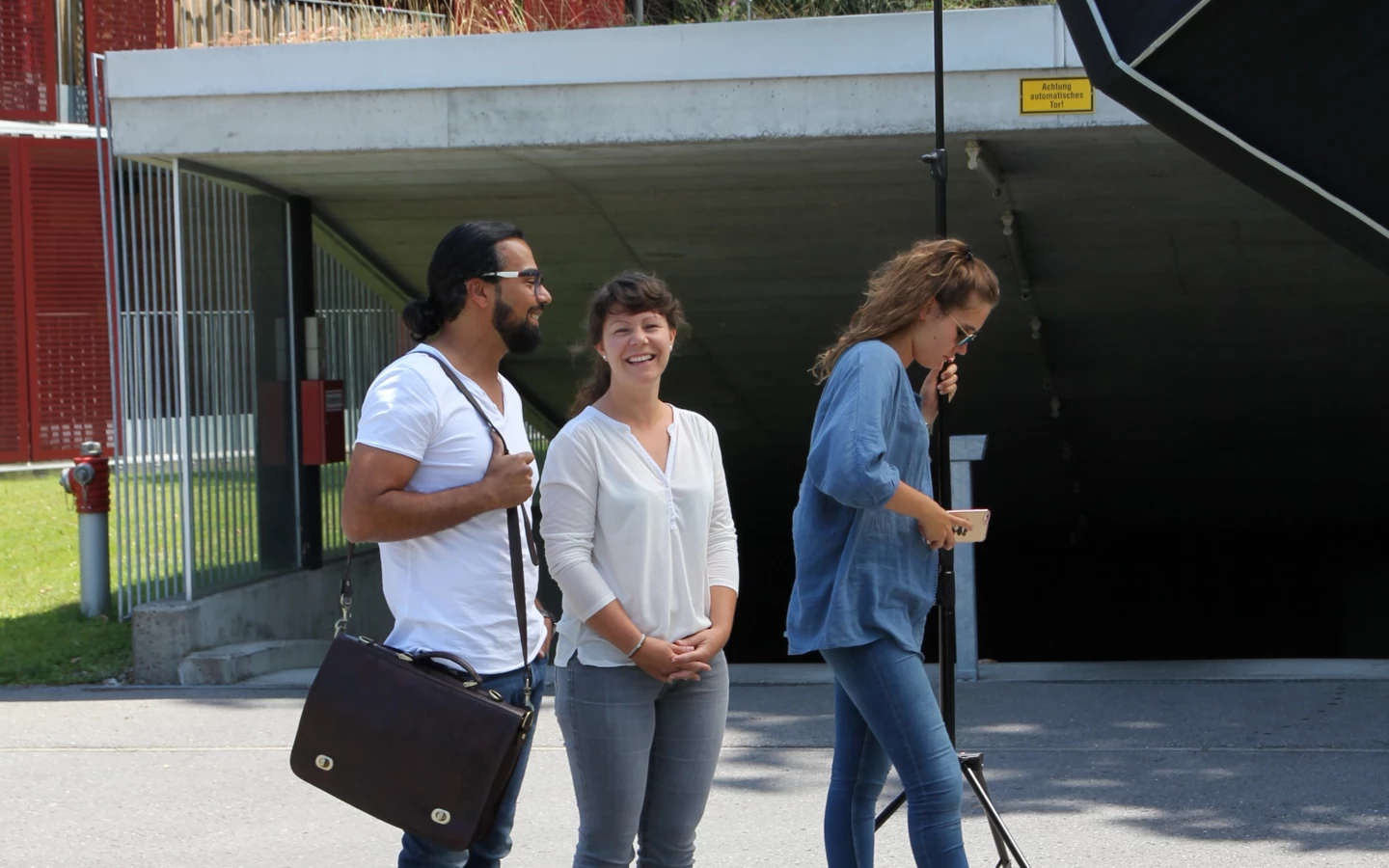 Zwei Frauen und ein Mann posieren lächelnd für ein Fotoshooting vor einem Gebäude mit roten Fassaden und einer Garageneinfahrt.