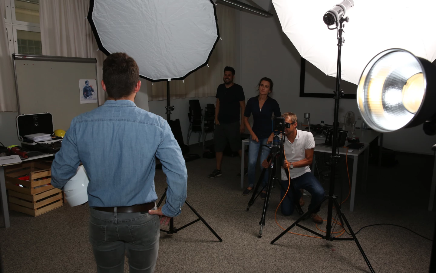 Ein Fotoshooting-Set mit einem Mann, der eine Bauarbeiter-Sicherheitskappe hat, vor Softboxen, während ein Kameramann filmt.