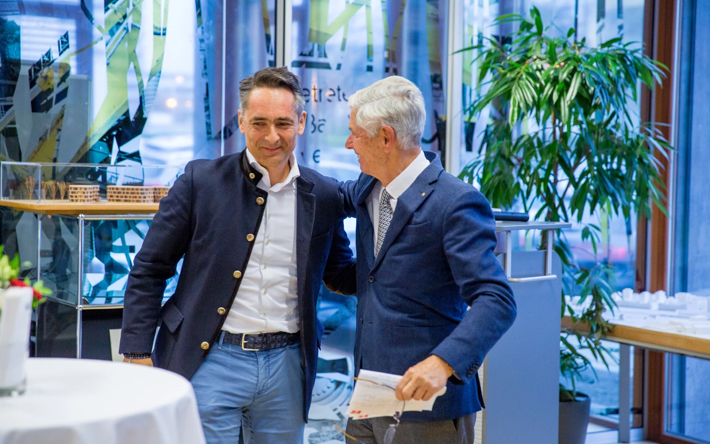 Zwei Männer in Anzügen unterhalten sich lachend bei einer Veranstaltung, mit Pflanzen und Architekturmodellen im Hintergrund (Hubert, Geburtstag).
