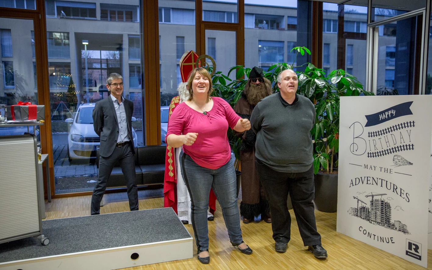 Vier Personen, darunter Nikolaus und Krampus, stehen im Innenbereich eines modernen Gebäudes neben einer großen Geburtstagskarte (Hubert).